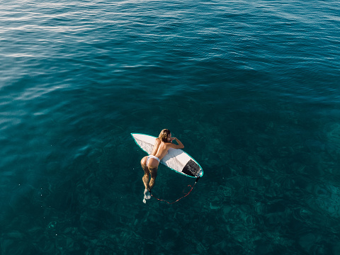 Sexy surf girl relaxing on surfboard in transparent ocean. Aerial view