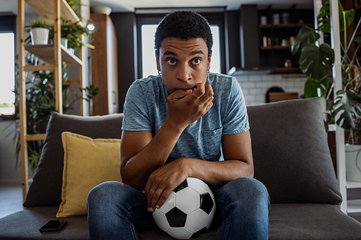 Portrait of young African American man watching a soccer game. Nervous man is sitting on the couch and biting fingernails while waiting the end of the game.