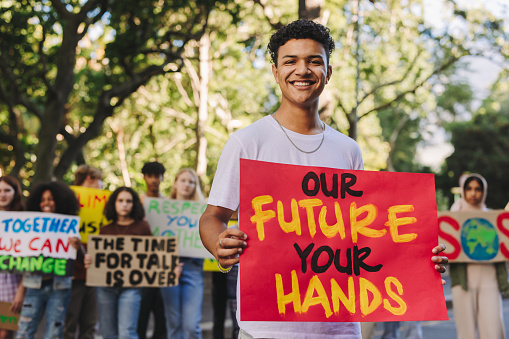 Happy teenage boy smiling at the camera while leading a march against climate change. Group of multiethnic youth activists protesting against global warming. Teenagers joining the global climate strike.