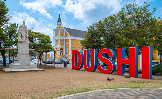 Big red blue word sign DUSHI in the city center of Willemstad, Curacao, Netherlands Antilles, Leeward Islands. Means beautiful and sweet, like the city. In the background a yellow church.