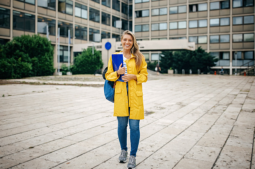 A young Caucasian woman in a yellow raincoat is smiling while holding a blue file folder.