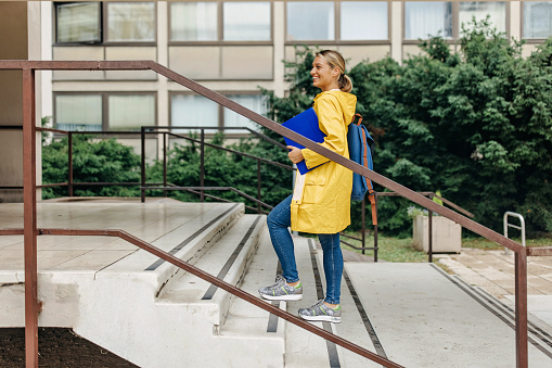 A young Caucasian woman in a bright yellow raincoat is walking up a flight of stairs, holding a blue file folder.