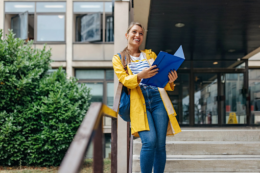 A young Caucasian woman with a beautiful smile is wearing a yellow raincoat and holding a blue file folder.