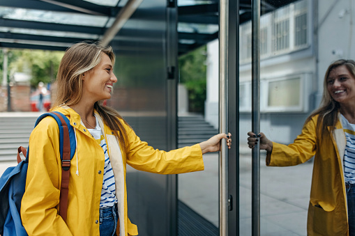A young Caucasian woman in a yellow raincoat with a big smile is opening a glass door.