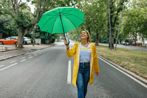 A young Caucasian woman is walking down the street in a yellow raincoat, carrying a green umbrella.