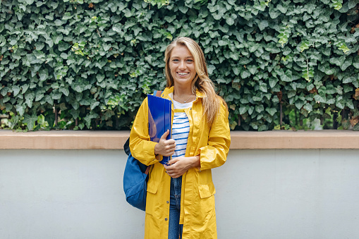 A young Caucasian woman is standing in front of a bush fence, wearing a yellow raincoat while smiling and looking at the camera.