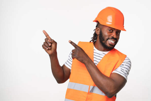 istock Young african-american man construction worker engineer architect in reflective orange vest and hardhat pointing at copy space showing free space isolated in white background 1405424120