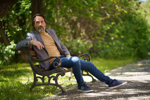 Mature freelancer man with grey beard, spend time sitting on the bench at the park enjoying free time or waiting on colleagues wearing casual jacket and jeans.