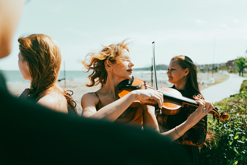 Three musicians in red dress and one musician in black suit at the beach. Music group playing violin, flute and double bass. Album cover.