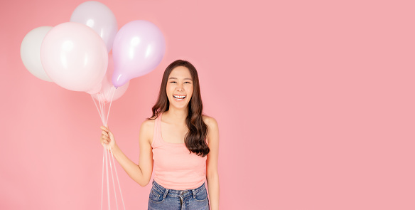 Happy pretty asian woman looking to camera in excitement holding pastel color balloon Expressive facial expression Cheerful girl hold balloons standing and laughing with happiness Pink background