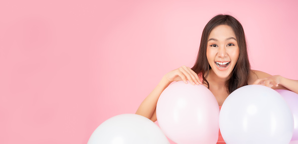 Happy cheerful asian woman looking at camera in excitement touching pastel color balloon Expressive facial expression Excited girl hiding behind balloons with happiness Pink background copy space