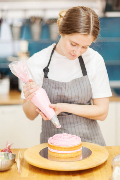 konditorin beim leckeren kuchen. frau, die rosa creme aus dem paspelbeutel auf eine biskuitschicht legt, die auf einem rotierenden kuchenständer in der bäckerei platziert wird - turn back stock-fotos und bilder