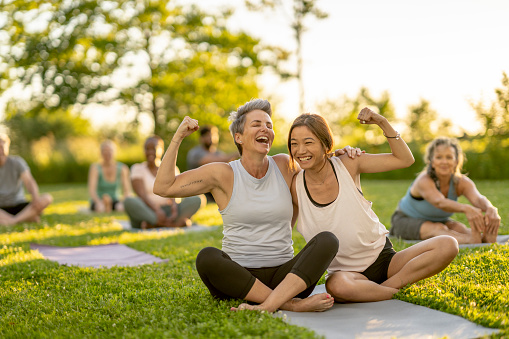 Two woman share a yoga mat as they lean in closely for a hug and flex their muscles.  They are seated outdoors among their peers during a group fitness class.  Each is dressed comfortably in athletic wear.