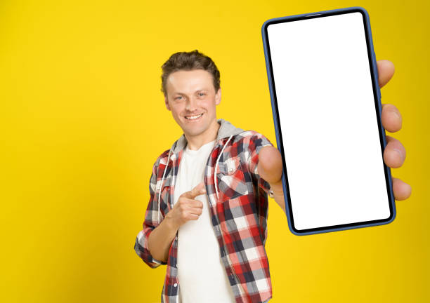 istock Happy to win handsome man pointing at smartphone with white empty screen, wearing red plaid shirt and jeans cellphone display mock up isolated on yellow background. Mobile app advertisement 1405434736
