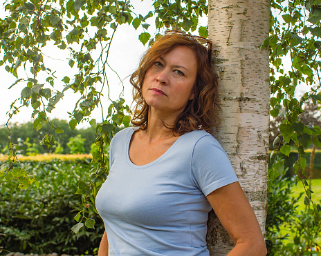 A mature woman stands near a birch tree with a sad look at the camera.