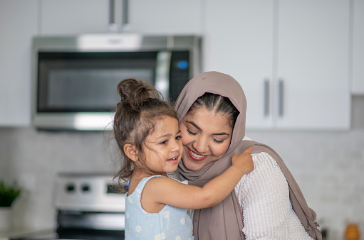 A Muslim Mother sits in the kitchen with her young daughter propped up in front of her, as they share a close moment.  They are both dressed casually and the mother is wearing a Hijab as they share an embrace.