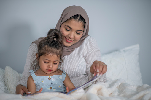 A Muslim Mother sits with her young daughter in bed as they read a book together.  They are both dressed casually and the mother is wearing Hijab as they share the story while snuggled in bed together.