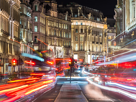 Red double decker buses rush between the spotlit shops and designer stores of Regent Street at night in the heart of London, UK.