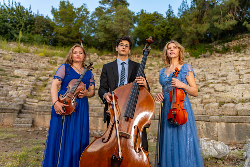 Two musicians in blue dress and one musician in black suit in ancient theater. Music group playing violin, flute and double bass. Album cover. Antalya, Kemer, Phaselis ancient city.