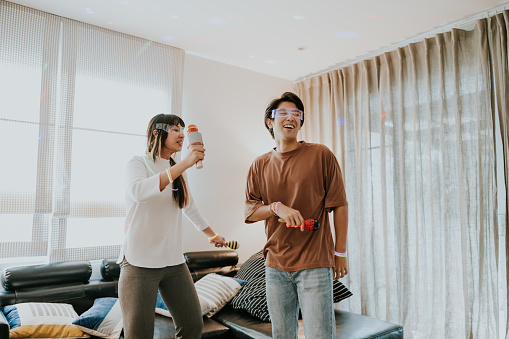 Asian heterosexual couple using microphone with karaoke time and dancing in front of the TV in their living room, simple living on weekend at home.