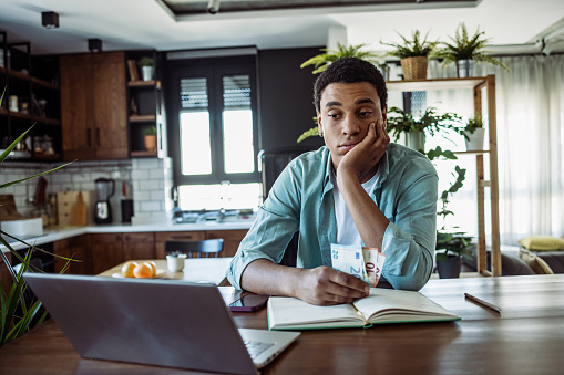 Portrait of young African American man sitting at the table in the kitchen and looking at the laptop. Desperate man is calculating his monthly expenses. Contemplation.