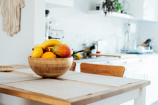 Home kitchen counter table with selective focus on bamboo bowl with exotic fruits on it with blurred background of modern cozy white kitchen. Home interior design details. Copy space