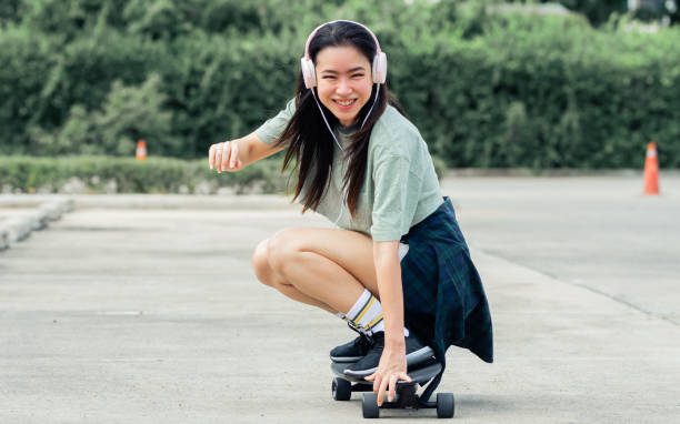 Beautiful sportive hipster happy Asian woman wearing casual shirt and shorts with headphone to listen music while playing skateboard as hobby in free time day on holiday, smiling at outdoor park. Beautiful sportive hipster happy Asian woman wearing casual shirt and shorts with headphone to listen music while playing skateboard as hobby in free time day on holiday, smiling at outdoor park Skateboarding stock pictures, royalty-free photos & images