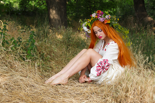 Beautiful slavic girl with long red hair with flower crown in white and red embroidered ethnic suit sitting on lawn. Feast of Ivan Kupala. Girl in national Ukrainian costume with wreath on her head