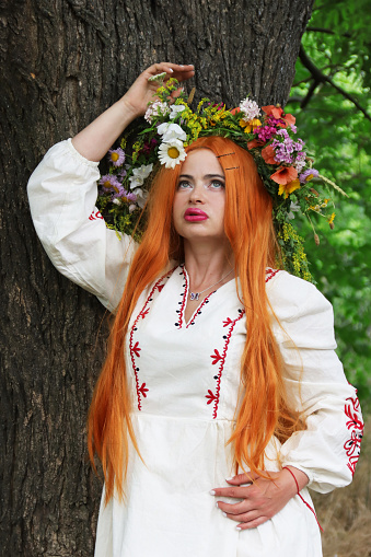 Beautiful slavic girl with long red hair with wreath from flowers in white and red embroidered ethnic suit stands by tree