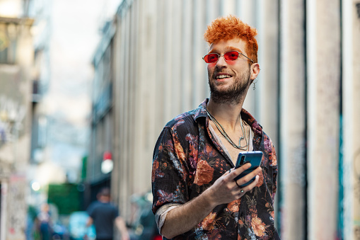 A Gay man standing on the street and smiling while holding smart phone.