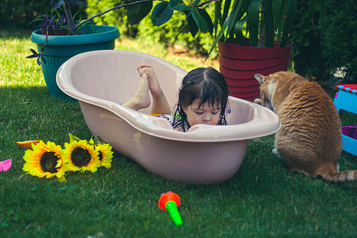 The child cools down herself in the tub, and the cat is sitting next to the tub.