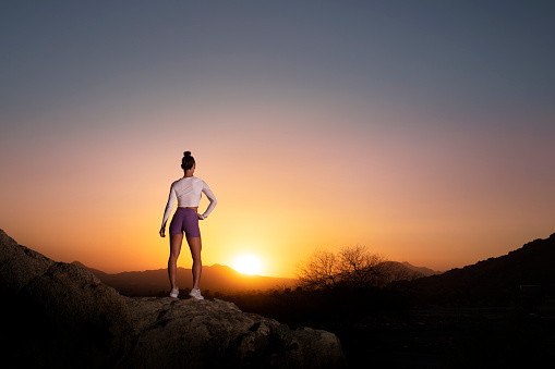 This is a conceptual photo showing a young fit woman standing on the rocks at sunset looking out at the horizon
