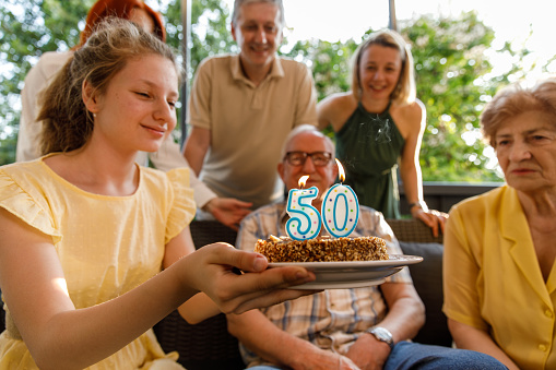 Selective focus shot of joyful teenage girl holding a cake with number 50 candles burning for her grandparents to blow out while celebrating their anniversary on the balcony with the rest of the family.