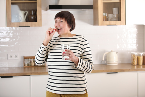 Happy middle aged 50s woman holding pill and glass of water taking dietary supplements. Portrait of smiling adult attractive woman taking care of health in menopause, at home