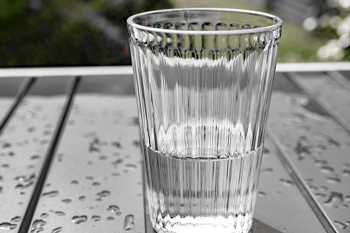 Glass of water, half full or half empty, resting on a metal garden table, still wet after a summer rain storm.  Belfast, Northern Ireland.