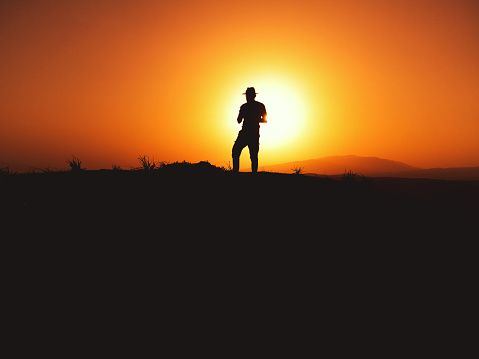 Silhouette of a man with fedora hat on sunset on a hill.