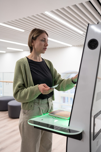 Middle-aged scandinavian woman using library self-service machine for borrow or return book, female holding ID card and clicking screen with finger, interacting with new smart library technologies