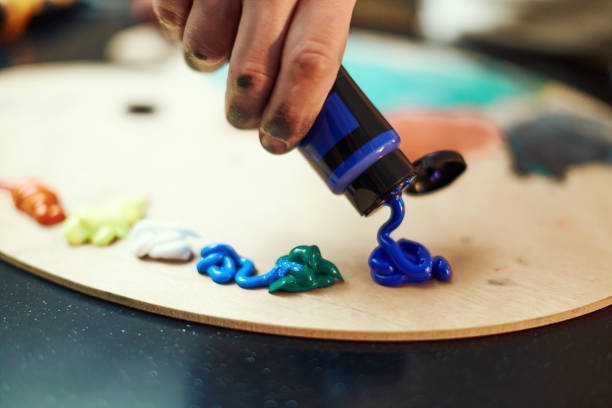 クローズアップ画家の手は、木製の絵画パレット上のチューブからアクリルブルーの塗料を絞ります。アーティストルーチン。クリエイティブな趣味 - アクリル画 ストックフォトと画像