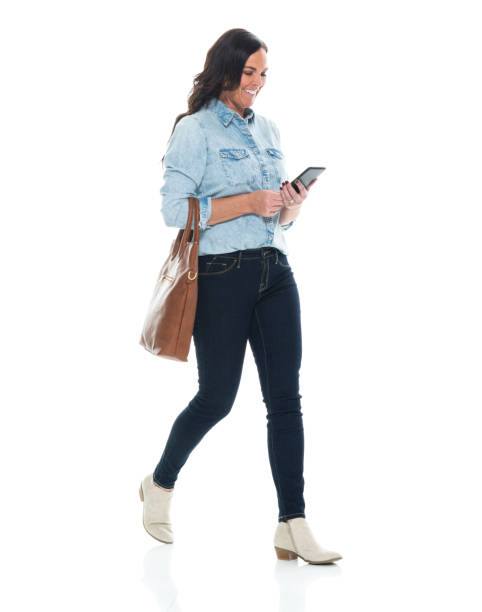 Caucasian young women walking in front of white background wearing jeans and holding purse and using smart phone Side view of aged 40-44 years old who is beautiful with brown hair caucasian young women walking in front of white background wearing jeans who is cheerful and holding purse and using smart phone older woman purse stock pictures, royalty-free photos & images