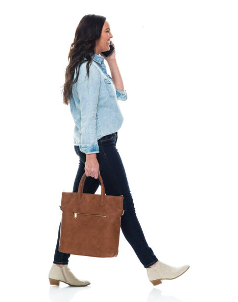 Caucasian female walking in front of white background wearing jeans and holding purse and using mobile phone Side view of aged 40-44 years old who is beautiful with long hair caucasian female walking in front of white background wearing jeans who is talking and holding purse and using mobile phone older woman purse stock pictures, royalty-free photos & images
