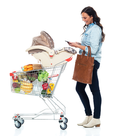 Profile view of aged 40-44 years old who is beautiful with brown hair baby - human age in front of white background wearing jeans who is feeling love who is shopping and holding car safety seat and using mobile phone