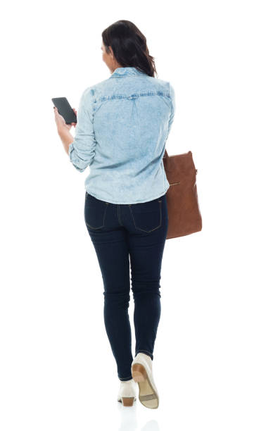 Caucasian young women walking in front of white background wearing button down shirt and holding purse and using smart phone stock photo