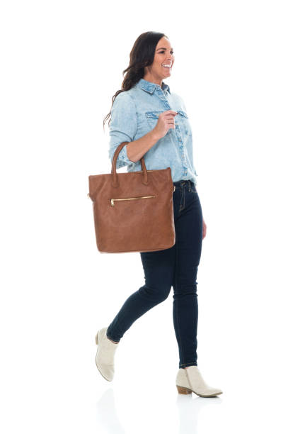 Caucasian young women walking in front of white background wearing button down shirt and holding purse Side view of aged 40-44 years old who is beautiful with brown hair caucasian young women walking in front of white background wearing button down shirt who is smiling and holding purse older woman purse stock pictures, royalty-free photos & images