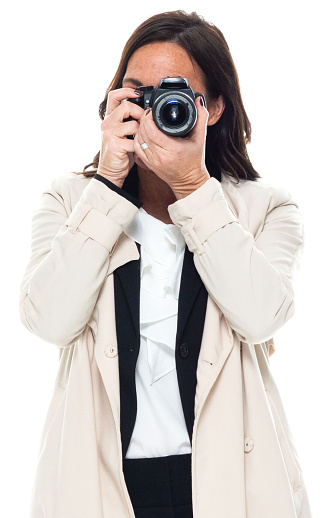 Front view of aged 40-44 years old who is beautiful with brown hair caucasian female tourist standing in front of white background wearing warm clothing who is happy who is photographing and holding camera