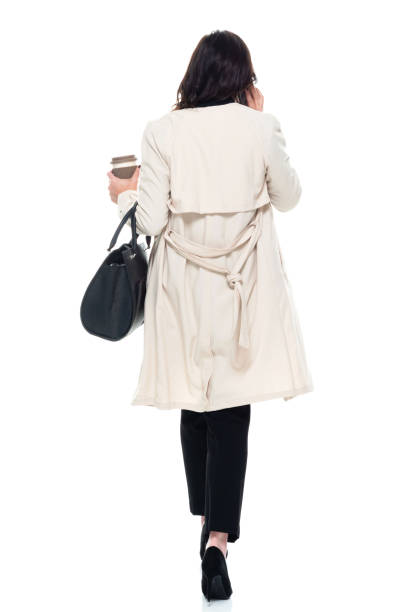 Caucasian young women businesswoman walking in front of white background wearing businesswear and holding purse and using mobile phone stock photo