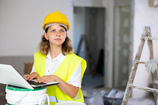 Young woman builder using laptop while working in apartment. Home improvement works.