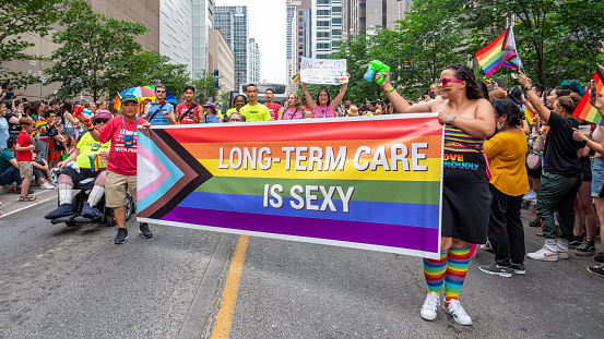 Toronto, Canada - June 26, 2022: A woman and a man are holding a rainbow flag banner as they walk in the Pride Parade on Bloor Street. Other people are seen walking and celebrating.