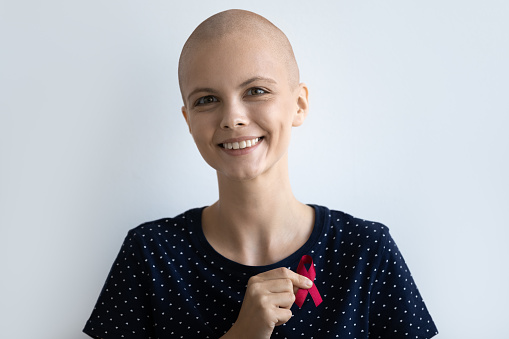 Happy beautiful millennial generation hairless after chemotherapy hopeful woman holding red pink ribbon in hands, showing support to cancer ill people, charity donation society awareness concept