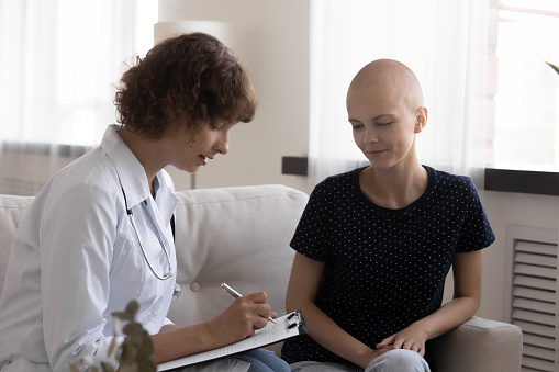 Concentrated young female doctor oncologist handwriting prescription or disease treatment to smiling millennial hairless after chemotherapy woman with cancer, doing regular checkup, healthcare concept
