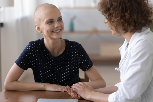 Smiling millennial generation bald after chemotherapy woman talking with professional female oncologist, listening to effective disease treatment results, consulting about cancer at checkup meeting.
